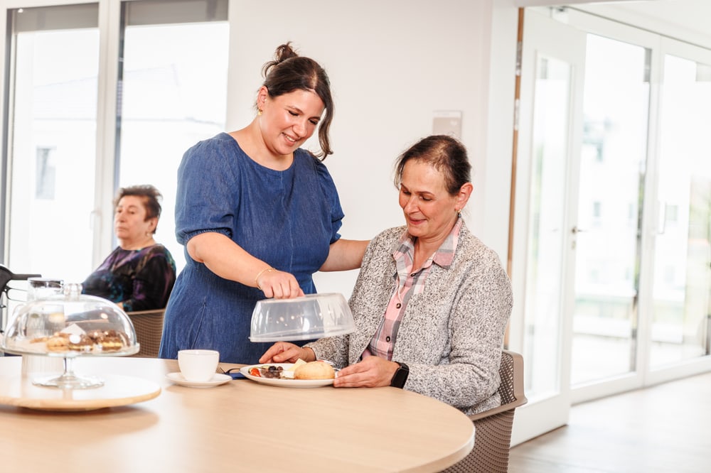 Pflegekraft serviert einer Seniorin Kuchen in der Tagespflege