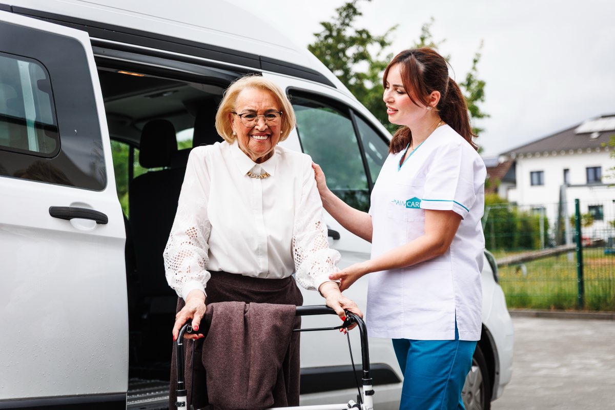 Pflegefachkraft begleitet eine Seniorin mit Rollator aus dem Main Care Fahrdienst